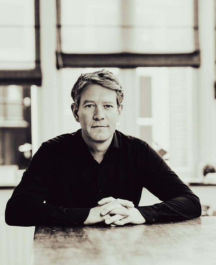 Portret van een man met lichtbruin haar, in zwart shirt, zittend aan een tafel met een neutrale achtergrond.