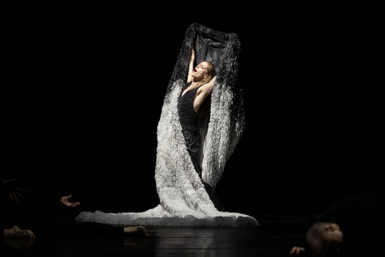 Een danseres gekleed in zwart, omringd door een witte doek, in een donkere theateromgeving.