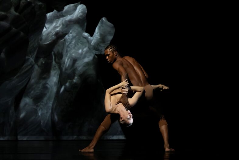 Twee dansers voeren een dynamische dans uit tegen een donkere achtergrond met abstracte projecties.