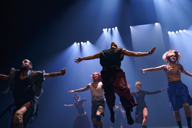 Zes dansers springen synchroon in het licht van een theater, met verschillende kostuums en expressieve houdingen.