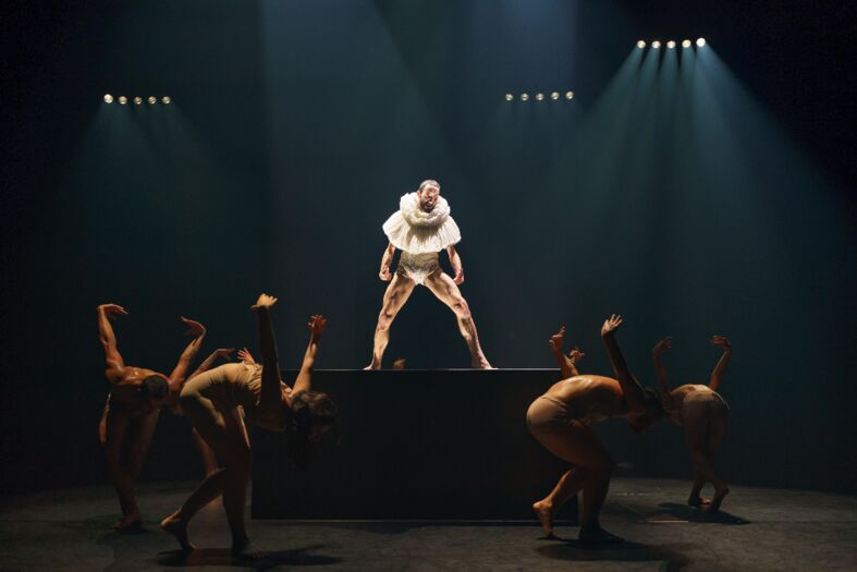 Dansers in een theatrale setting, met een centrale performer op een verhoging, belicht door spots.