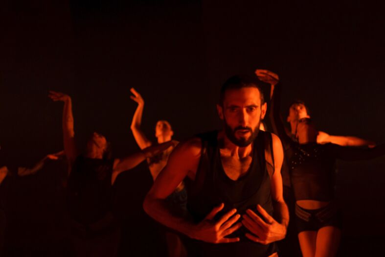 Een groep dansers in donkere kleding voert een energieke dansuitvoering uit in een gedimd, oranje verlichte ruimte.