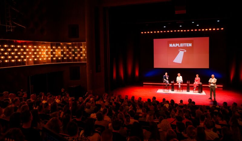 Een volledig gevulde theaterzaal met een podium waarop vier mensen zitten, terwijl een spreker het publiek toespreekt.