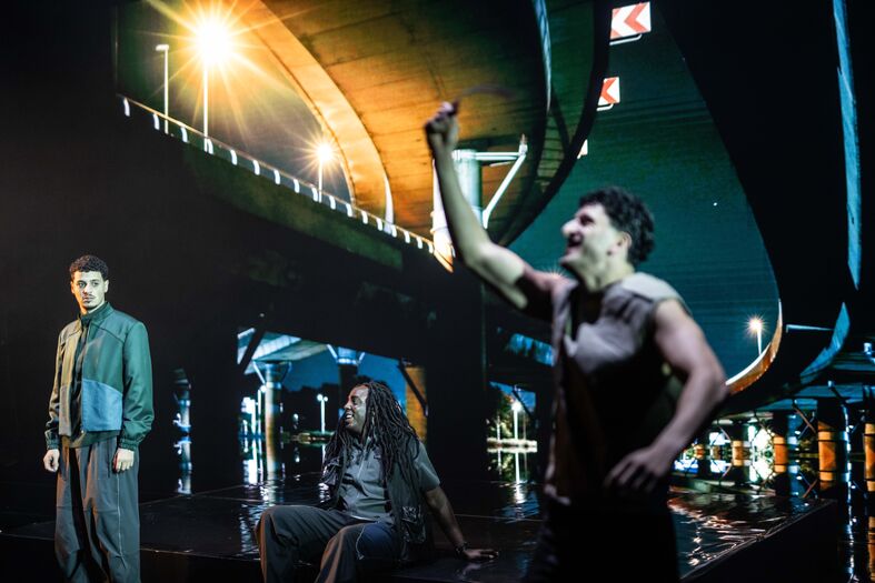 Theater voorstelling onder een viaduct met drie acteurs in verschillende poses, verlicht door kunstlicht.