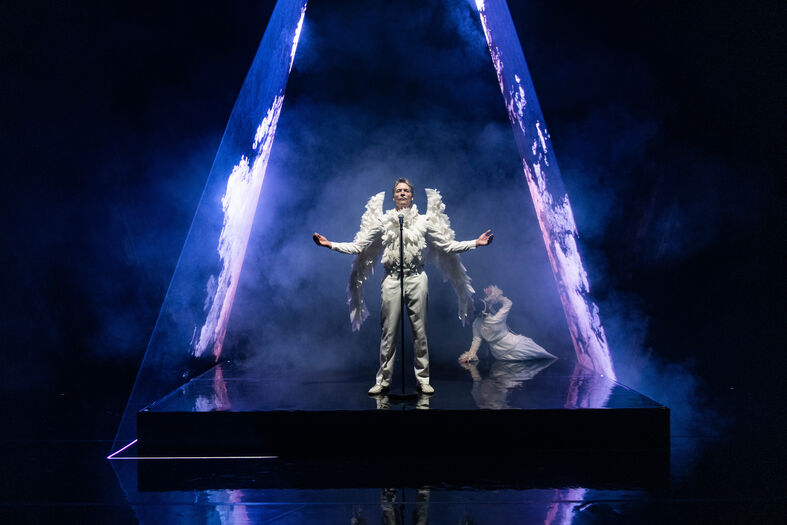 Optreden in een pyramidevormig podium met een zanger in witte kleding en vleugels, omringd door rook en gekleurde lichten.