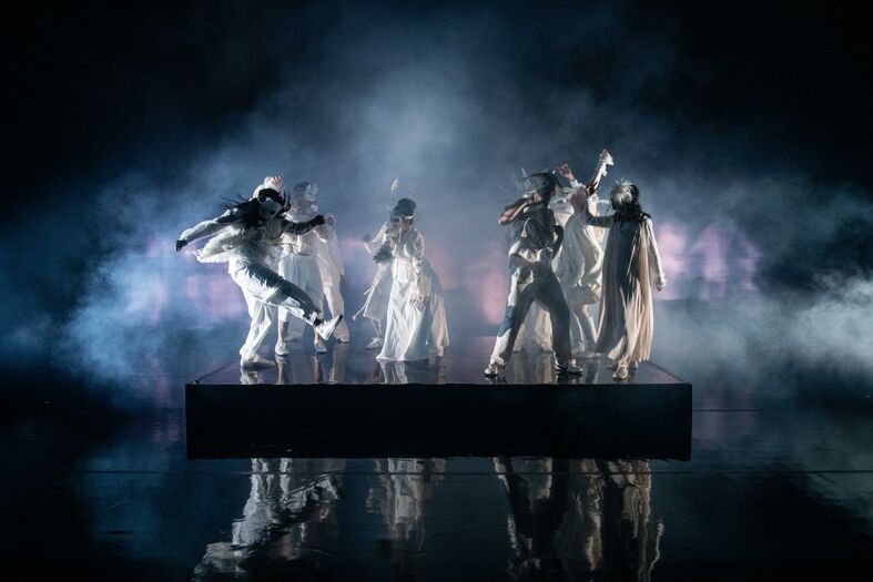 Toneelvoorstelling met zes acteurs in dramatische pose, gekleed in witte kostuums, omgeven door mist en lichteffecten.