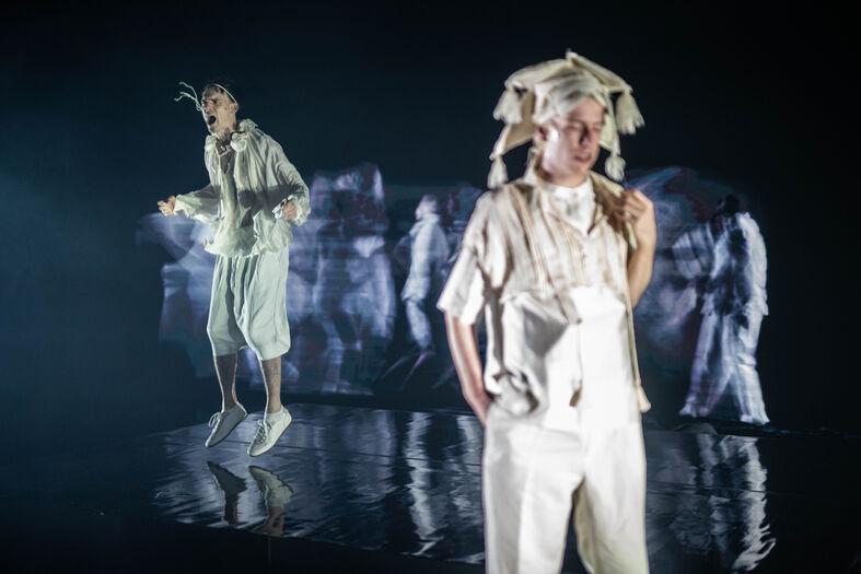 Twee performers in creatieve kostuums staan op een donkere podium met een abstracte achtergrond.