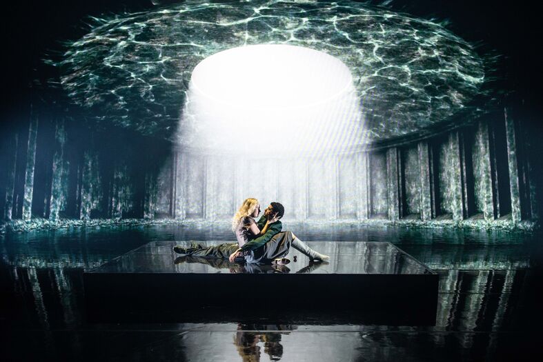Een man en vrouw zitten samen in een spiegelachtige ruimte met water, met een lichtbundel die vanuit het plafond valt.