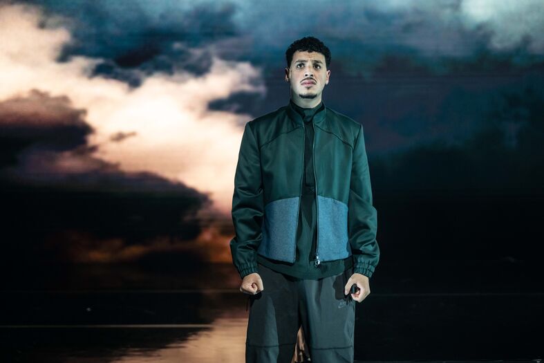 Man in een zwarte en grijze outfit staat op een podium met een dramatische achtergrond van donkere, bewolkte lucht.
