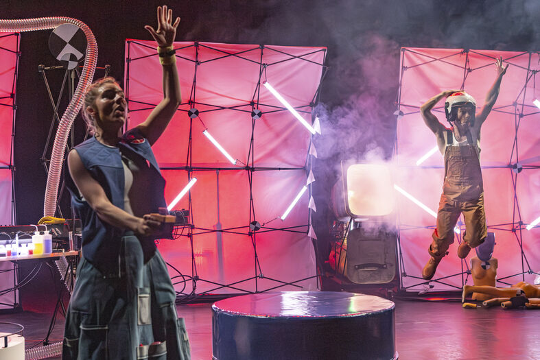 Twee performers op een podium met kleurrijke lichten en rook, een van hen zwaait terwijl de ander in een kostuum staat.