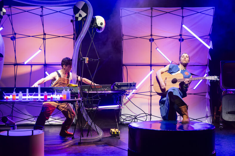 Twee muzikanten op een podium, één speelt gitaar terwijl de ander een synthesizer bedient, omgeven door kleurrijke lichten.