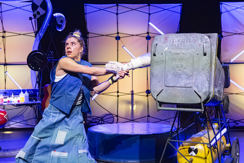 Een actrice in een creatief kostuum slaat met een plastic voorwerp op een grote metalen container op een podium.