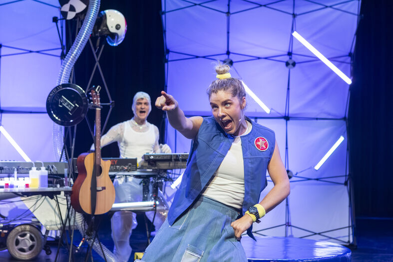 Twee performers op het podium: een met een gitaar en de ander in actieve pose met een kleurrijke outfit en gelach.