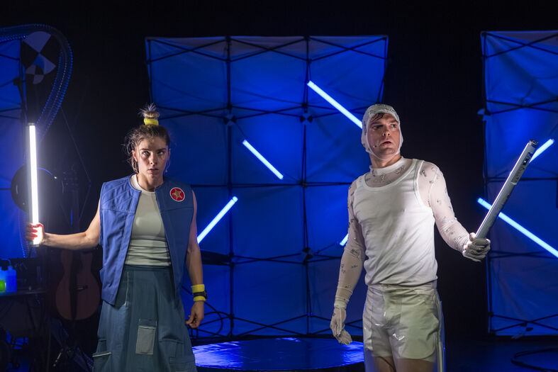 Twee performers op het podium, één met een lichtstaaf en de ander in een futuristische outfit, tegen een blauwe lichtachtergrond.