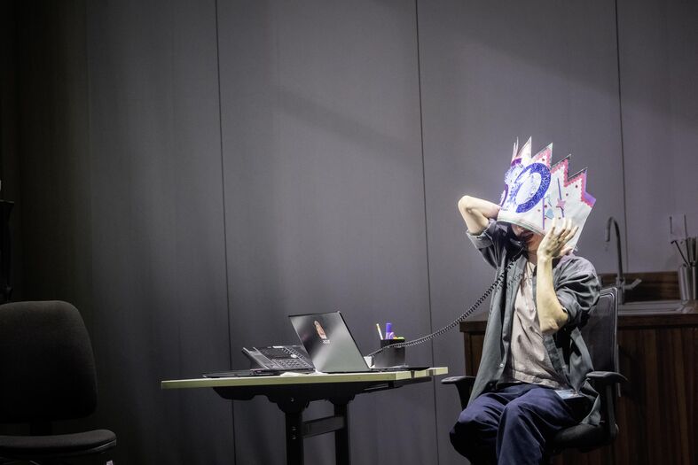 Een persoon met een kleurrijke hoofdtooi zit aan een tafel met een laptop en papieren in een zaal.