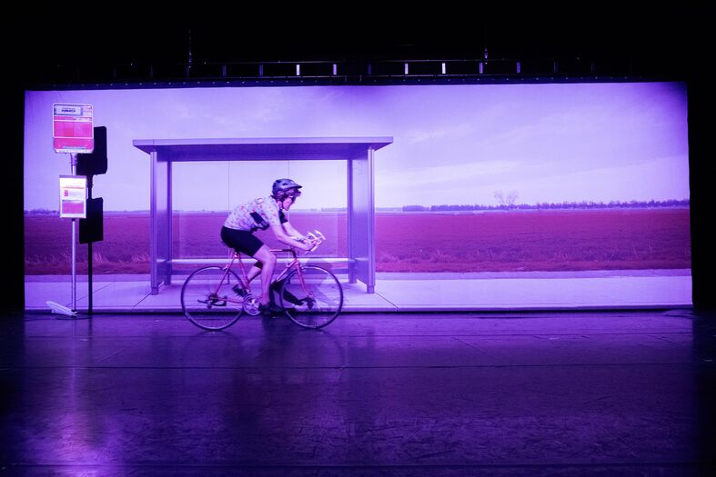 Fietser op een monitor met een paarse achtergrond, voor een bushalte rode verkeerslichten.