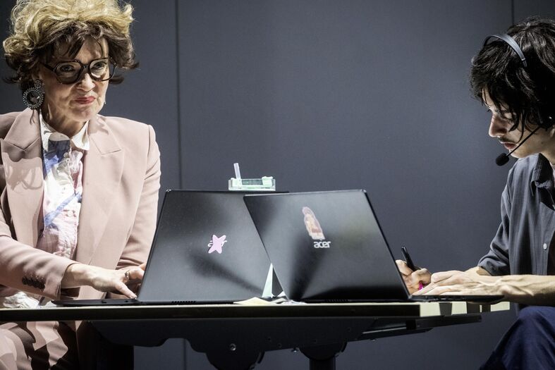 Twee vrouwen in zakelijke kleding zitten aan een tafel met laptops, geconcentreerd bezig met een gesprek of presentatie.
