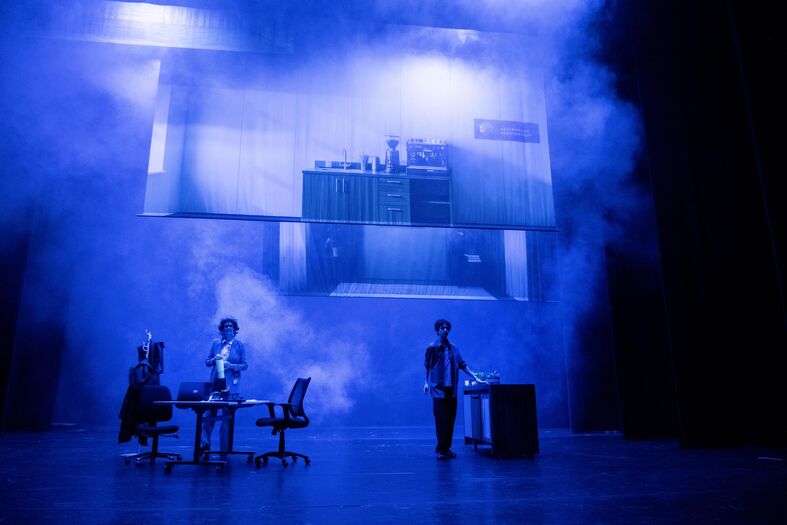 Twee acteurs op een podium met blauwe verlichting, omringd door rook en een groot scherm op de achtergrond.