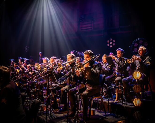 Orkest van muzikanten die trompetten spelen op een podium, verlicht door sfeervolle verlichting.