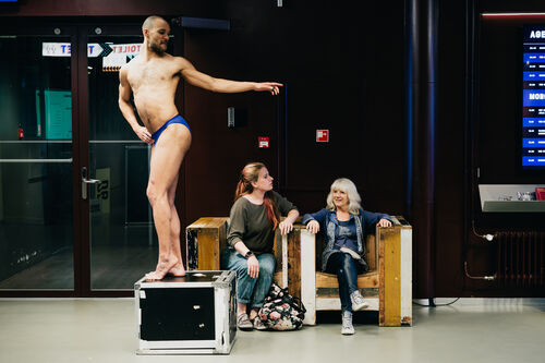 Man in zwembadoutfit poses op een kist, terwijl twee vrouwen op een bankje kijken en lachen in een openbare ruimte.
