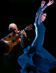 Een man met gitaar speelt terwijl een vrouw in een blauwe jurk danst, tegen een donkere achtergrond.
