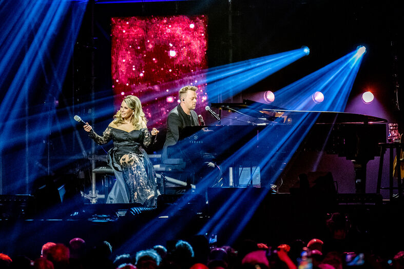 Zangeres en pianist optreden op een podium met kleurrijke verlichting en een abstracte achtergrond.