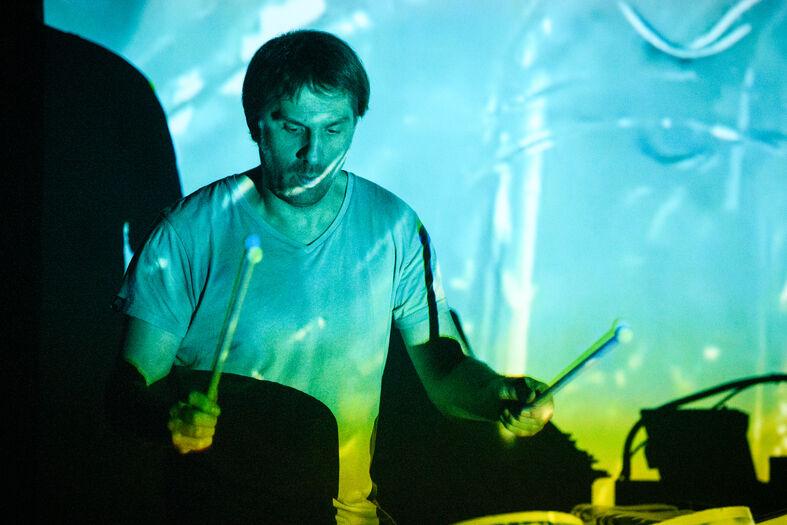 Drummer met stokken, verlicht door kleurrijke projectorlicht, terwijl hij speelt in een donkere omgeving.