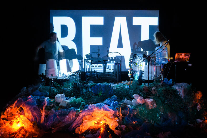 Twee muzikanten op een podium met een groot scherm waarop het woord "BEAT" staat afgebeeld, omringd door kleurrijke belichting.