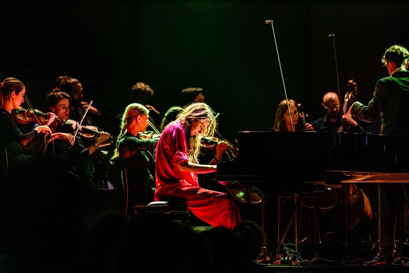 Een vrouwelijke pianist speelt op een vleugelpiano, omringd door een orkest in een sfeervolle, donkere setting.