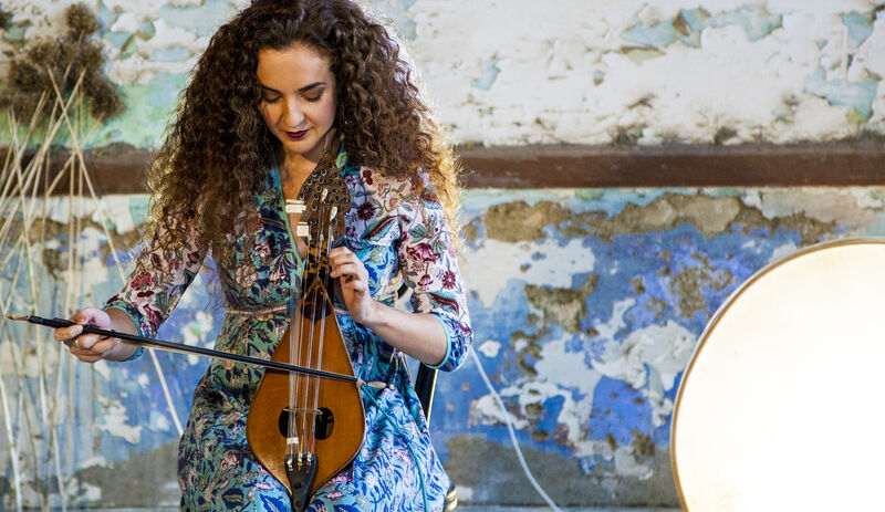 Een vrouw met krullend haar speelt een snaarinstrument in een kleurrijke ruïne met een grote lamp op de achtergrond.