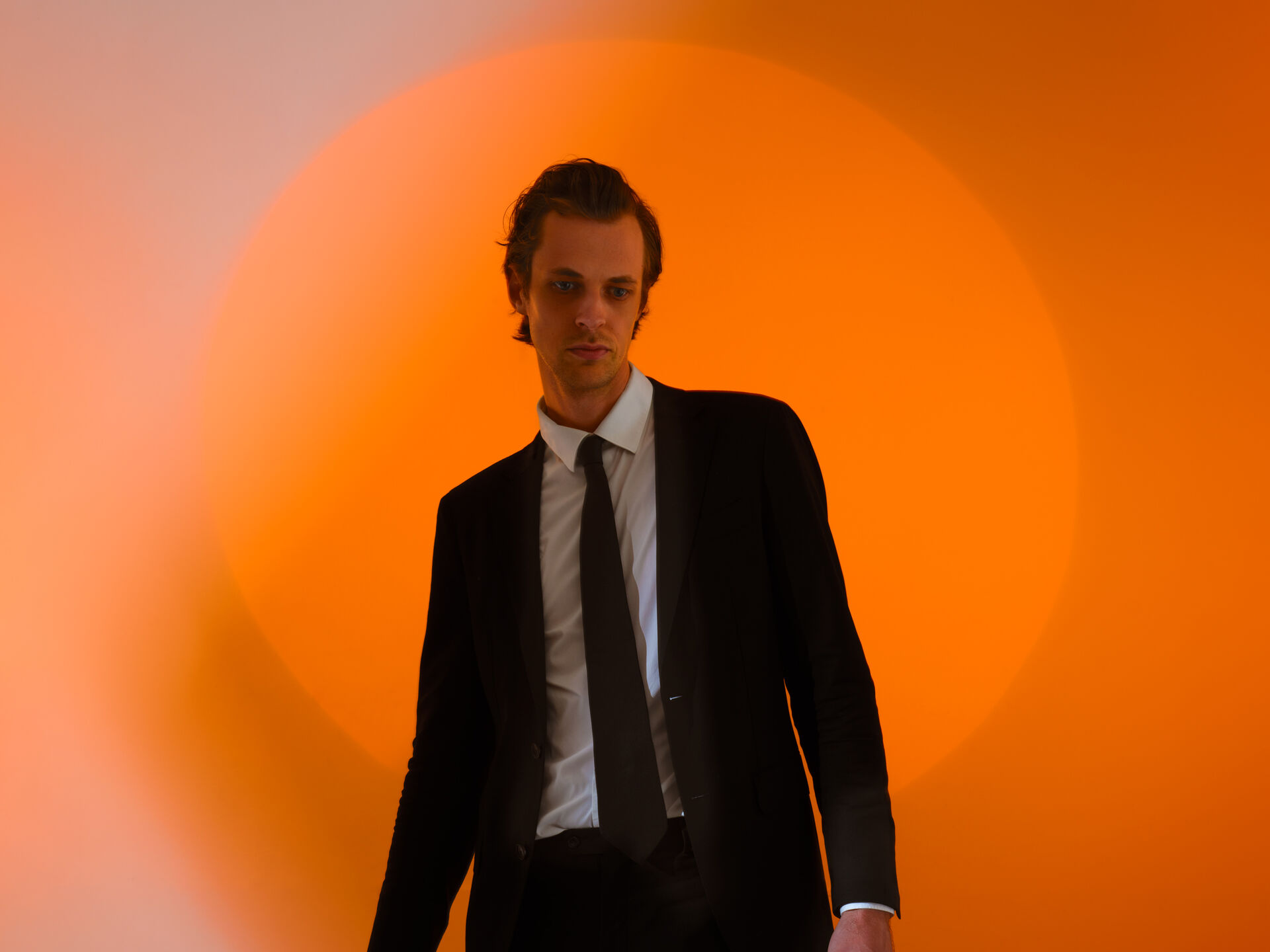 Persoon (David Linszen) Persoon in donker pak staat in zachte spotlight voor een grote cirkel van warm oranje licht.