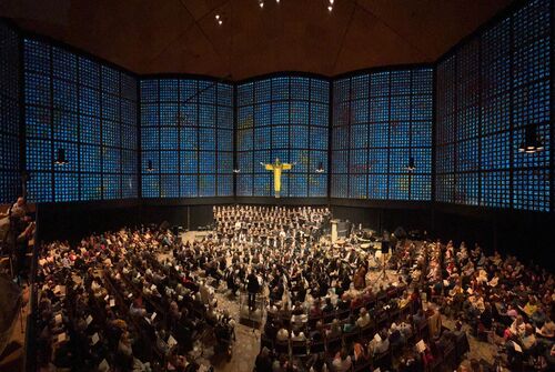 Een grote zaal met een koor en orkest, omringd door glazen wanden en een kruis aan de muur.