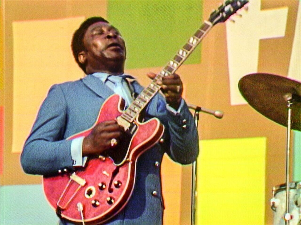 B.B. King speelt gitaar op een podium, gekleed in een blauw pak, met een drummer op de achtergrond.