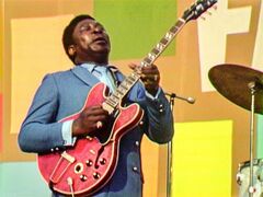 B.B. King speelt gitaar op een podium, gekleed in een blauw pak, met een drummer op de achtergrond.