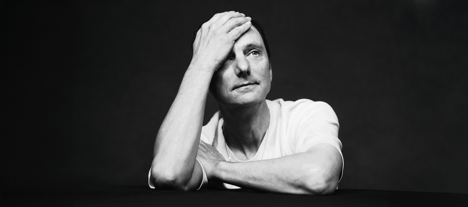Thomas van Luyn in wit T‑shirt leunt voorover met één hand op het voorhoofd, in een zwart‑wit portret tegen donkere achtergrond.