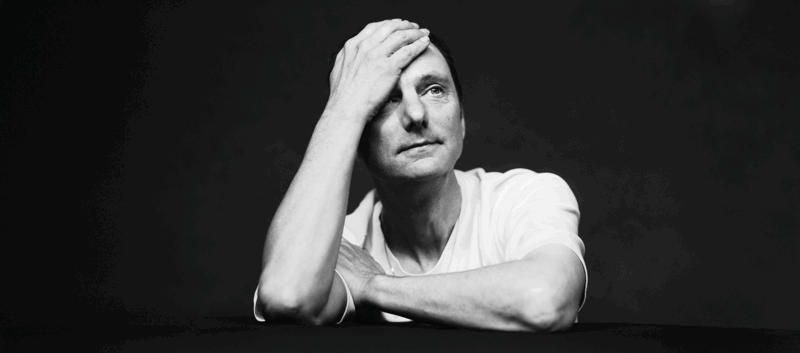 Thomas van Luyn in wit T‑shirt leunt voorover met één hand op het voorhoofd, in een zwart‑wit portret tegen donkere achtergrond.