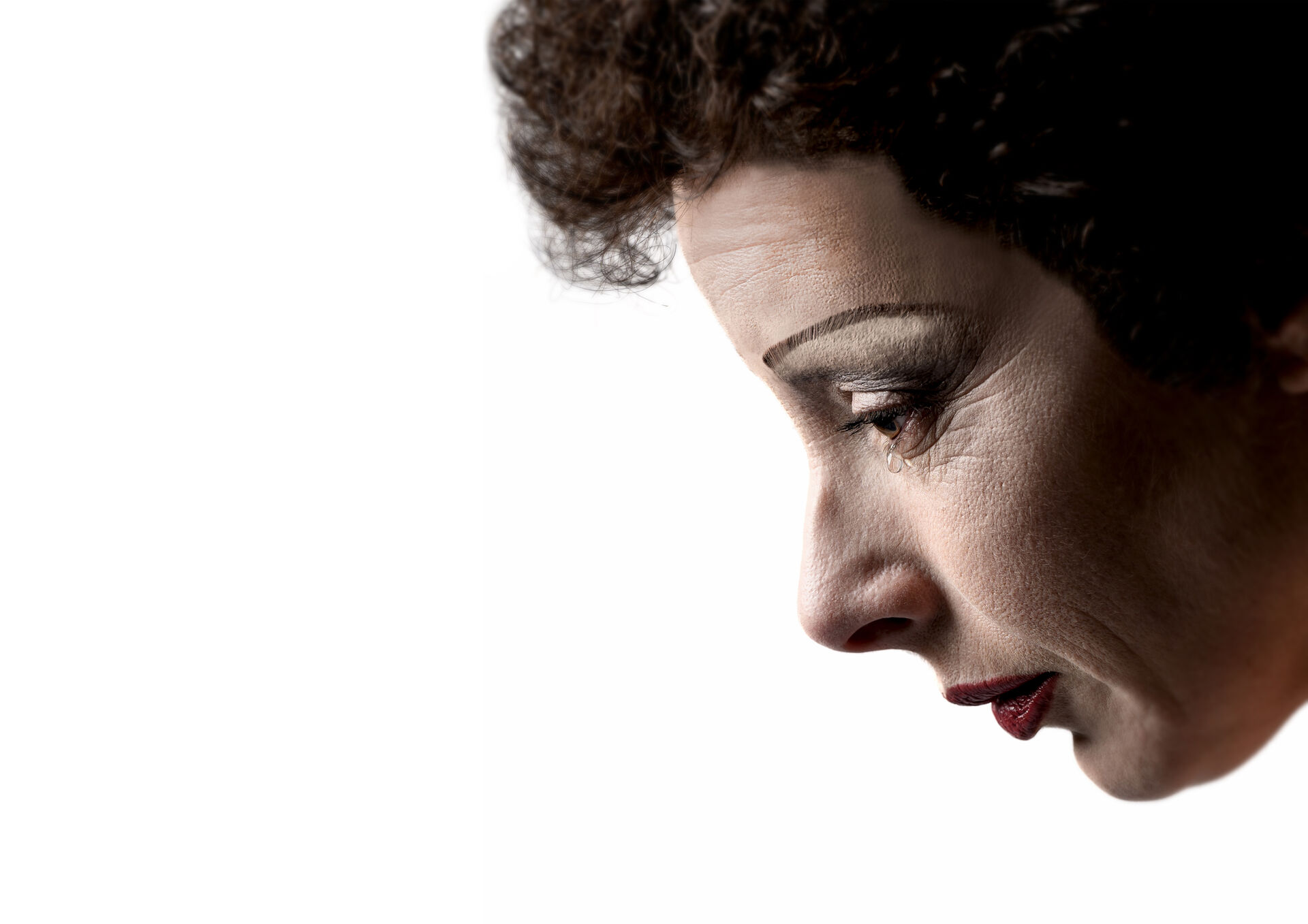 Zijaanzicht van een persoon (Piaf) met donkere krullen en make-up, met een traan langs het oog op een witte achtergrond.