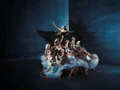 Groep balletdansers in witte tutu’s ondersteunt centrale figuur met zwarte vleugels en uitgestrekte armen.