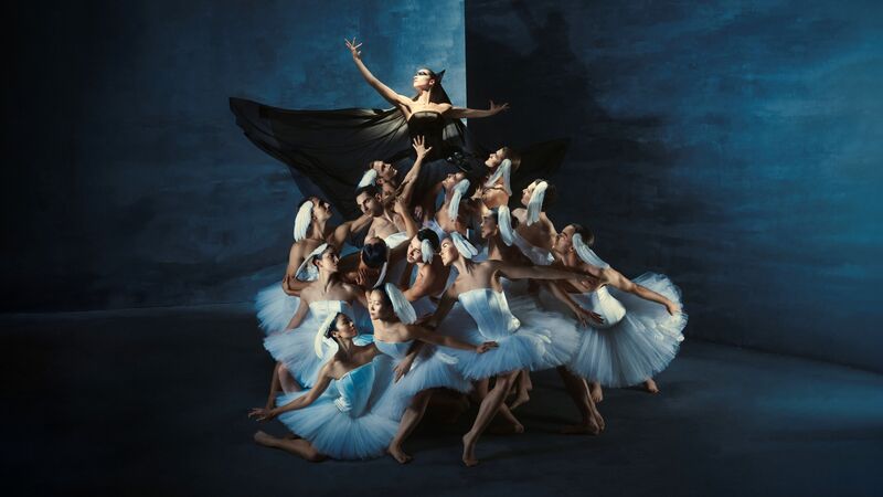 Groep balletdansers in witte tutu’s ondersteunt centrale figuur met zwarte vleugels en uitgestrekte armen.