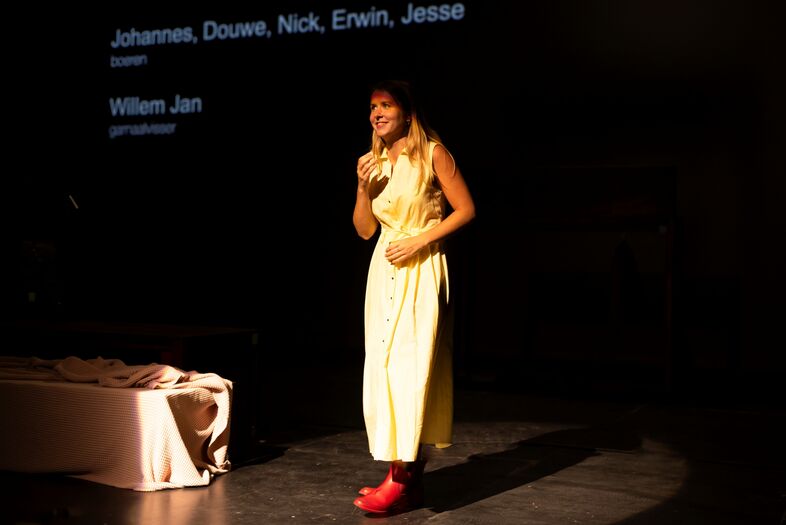Vrouw in een gele jurk en rode schoenen staat op een podium, spreekt tot het publiek met een microfoon in de hand.