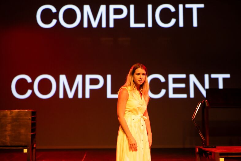 Een actrice in een witte jurk staat op een podium met de woorden "COMPLICIT" en "COMPLACENT" achter haar geprojecteerd.