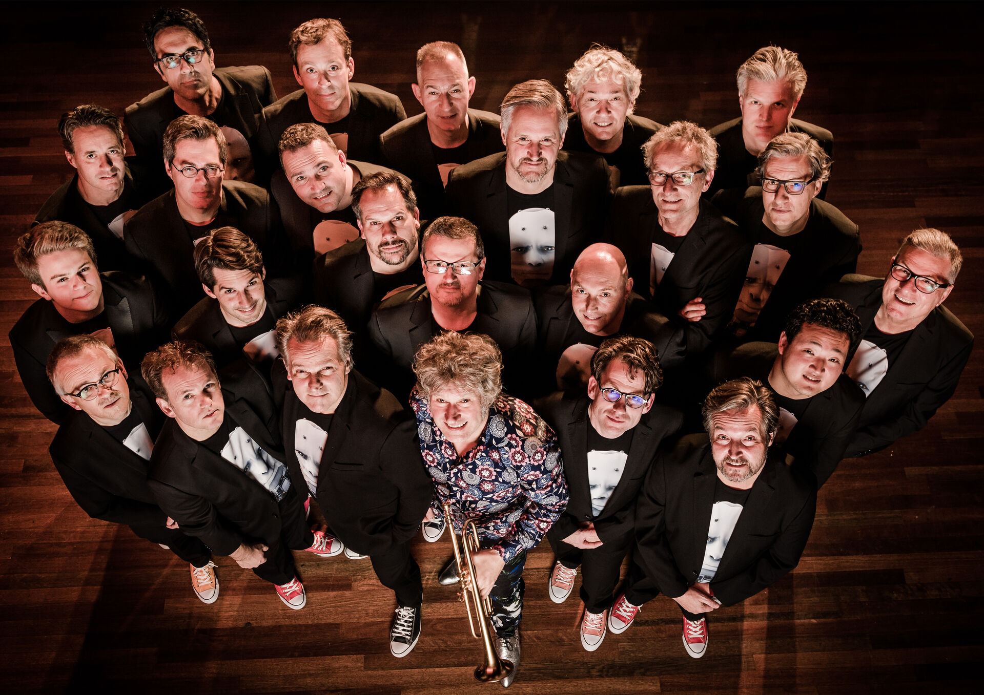 Grote groep muzikanten in zwarte kleding staat samengedrongen op houten vloer, één persoon met trompet centraal van bovenaf gefotografeerd.