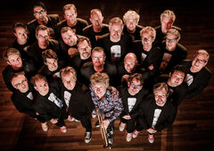 Grote groep muzikanten in zwarte kleding staat samengedrongen op houten vloer, één persoon met trompet centraal van bovenaf gefotografeerd.