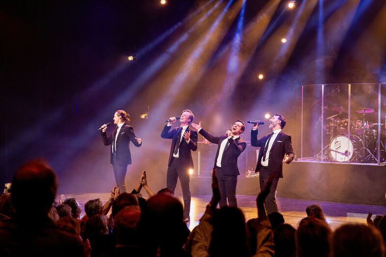 Vier zangers in zwarte pakken performen op een podium, omringd door een enthousiast publiek en sfeervolle verlichting.