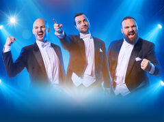 Drie mannen in formele kleding staan op een podium, enthousiast wijzend en juigend onder blauwe lichtstralen.