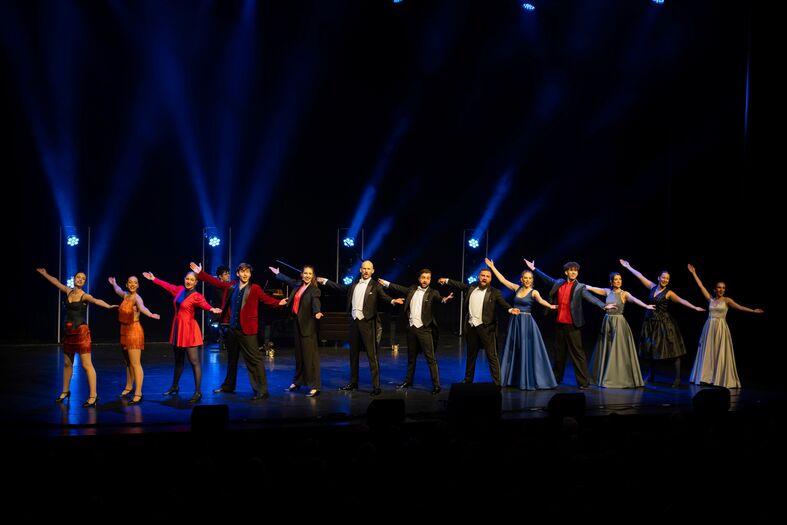 Groep performers in avondkleding, op een podium, met uitgestoken armen en een kleurrijke lichtshow op de achtergrond.