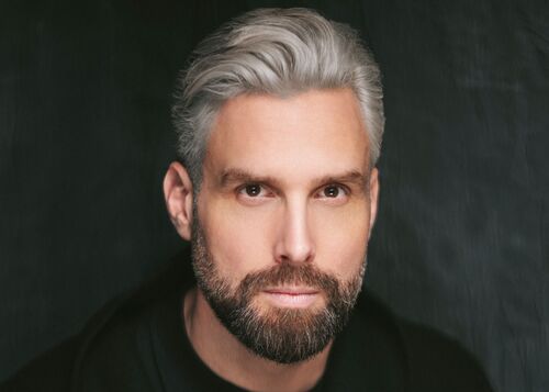 Persoon (Nick Schilder) met zilvergrijs haar en baard kijkt recht in camera.