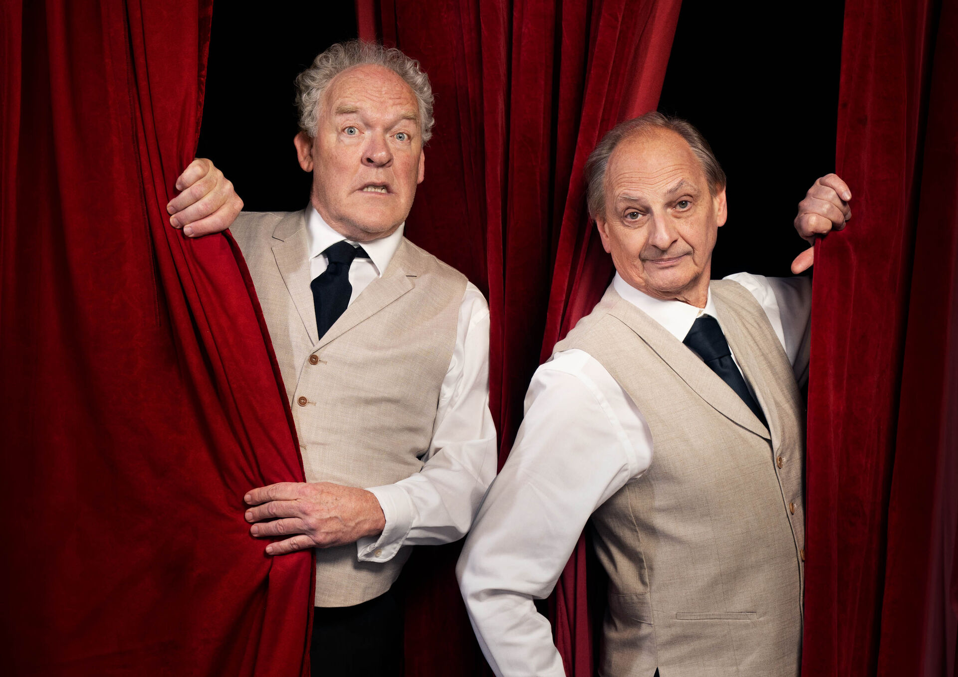 Bert Visscher & Martin van Waardenberg  in tweedelige kostuums staan achter een rode theatergordijn en kijken verbaasd naar de kamera.