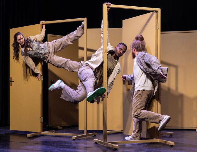 Drie performers hangen en springen dynamisch aan gele deuromlijstingen tijdens een choreografie
