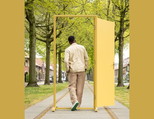 Persoon loopt door een vrijstaande gele deur op een met bomen omzoomd wandelpad.